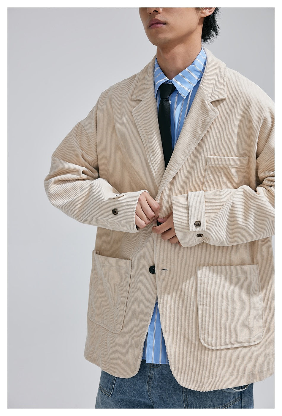 Close view of a model wearing a cream corduroy blazer with three patch pockets over a blue striped shirt and black tie.