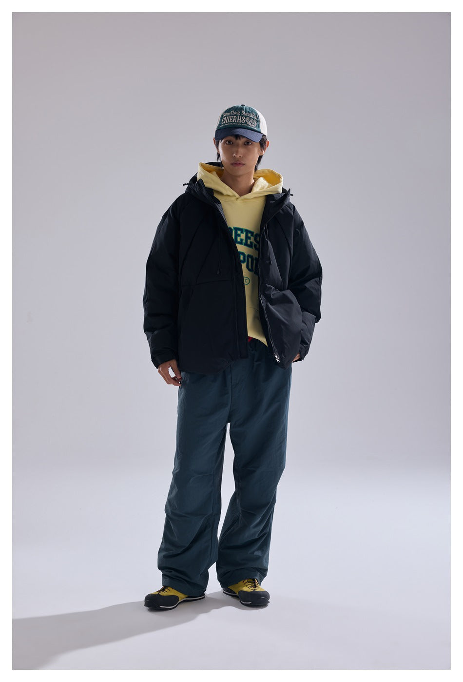 Model wearing an open black hooded puffer jacket over a yellow hoodie with teal pants and a cap, full-length view