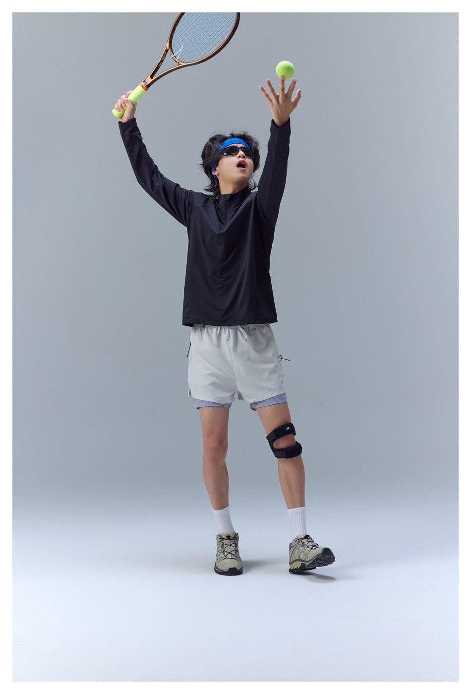 Model wearing a black long-sleeve quarter-zip top and light shorts, tossing a tennis ball with racket raised.