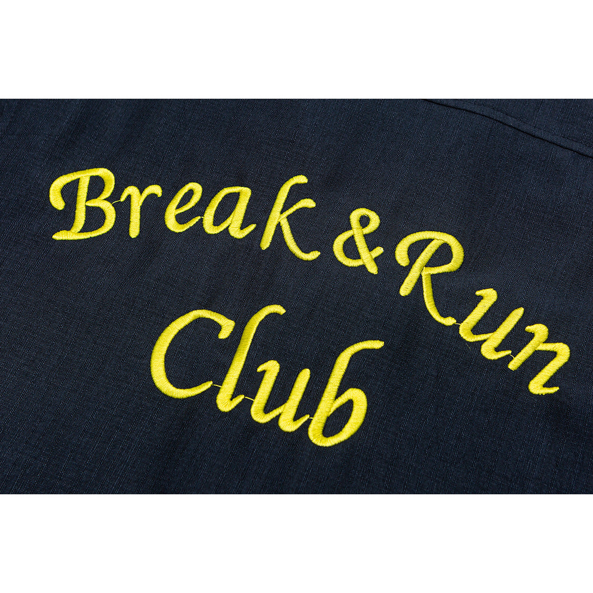 Close-up of yellow “Break & Run Club” embroidery on dark navy shirt fabric.