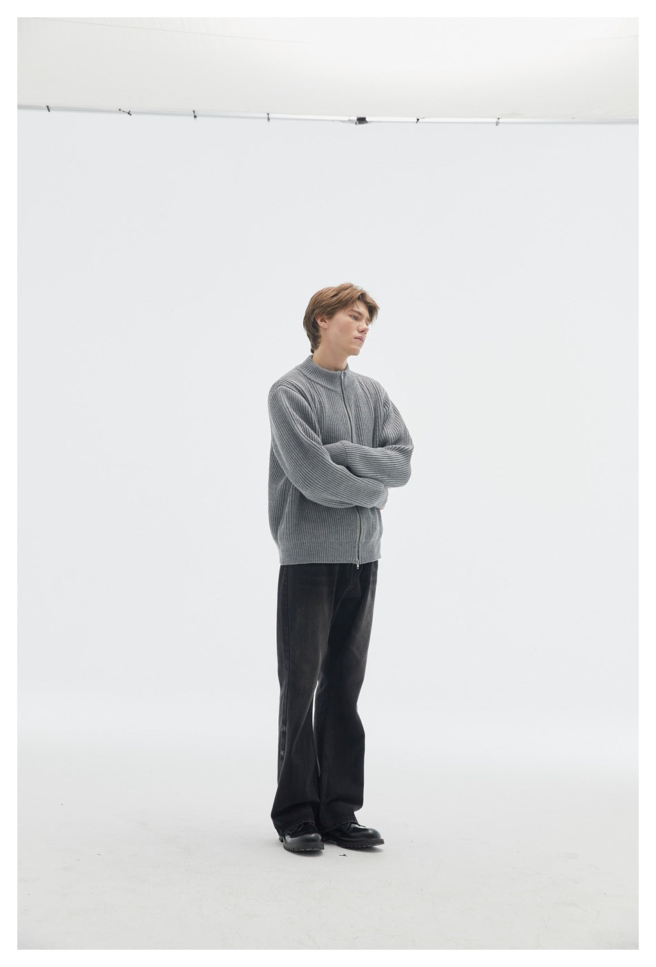Side view of a model wearing a gray ribbed zip cardigan and black trousers, arms crossed.
