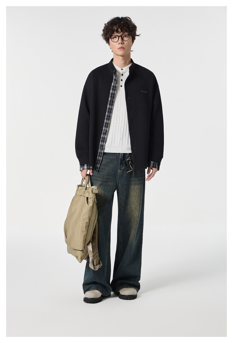 Model wearing the black jacket open over a white knit top with plaid placket showing, wide jeans, holding a tan bag