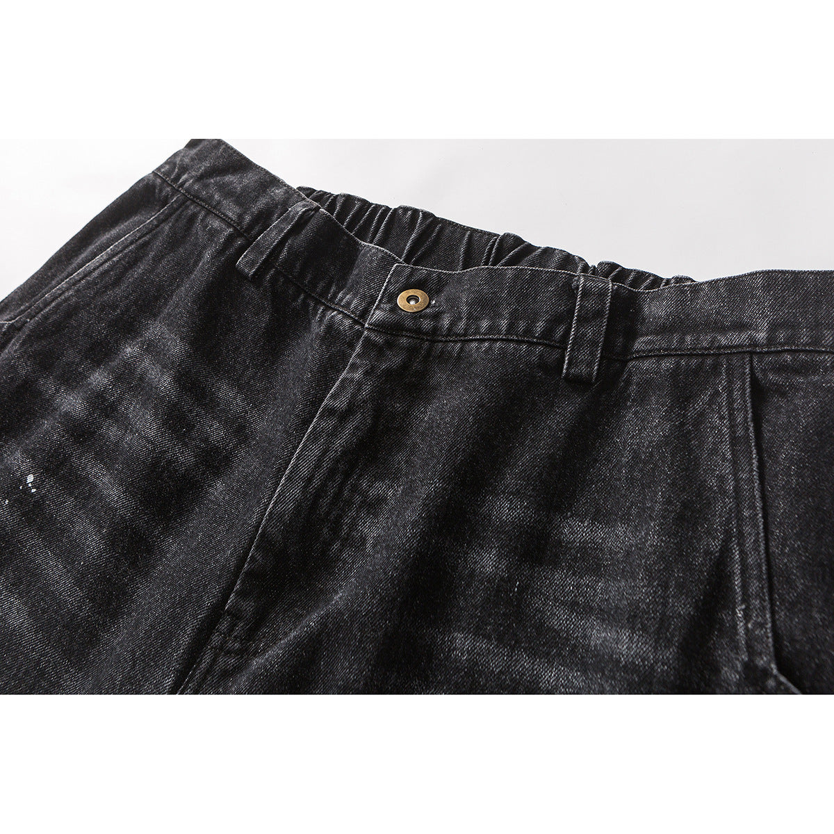 Close-up of black washed trousers showing the waistband, belt loops, brass button, and subtle white paint flecks.