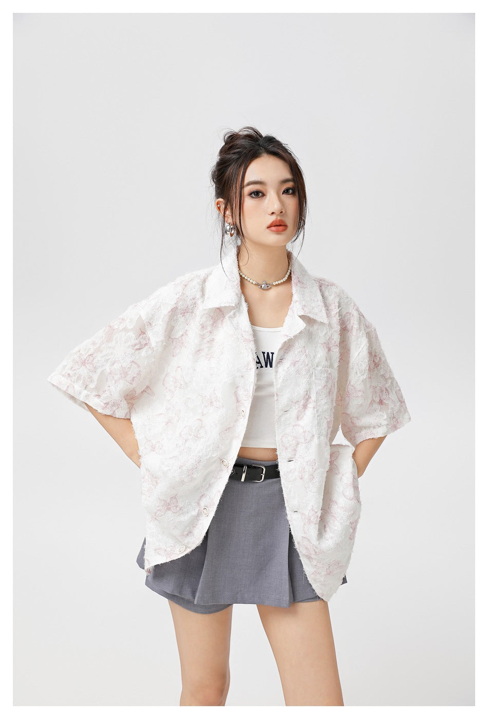 Model in an oversized white short-sleeve shirt with pink floral texture, open over a white tank and gray pleated skirt