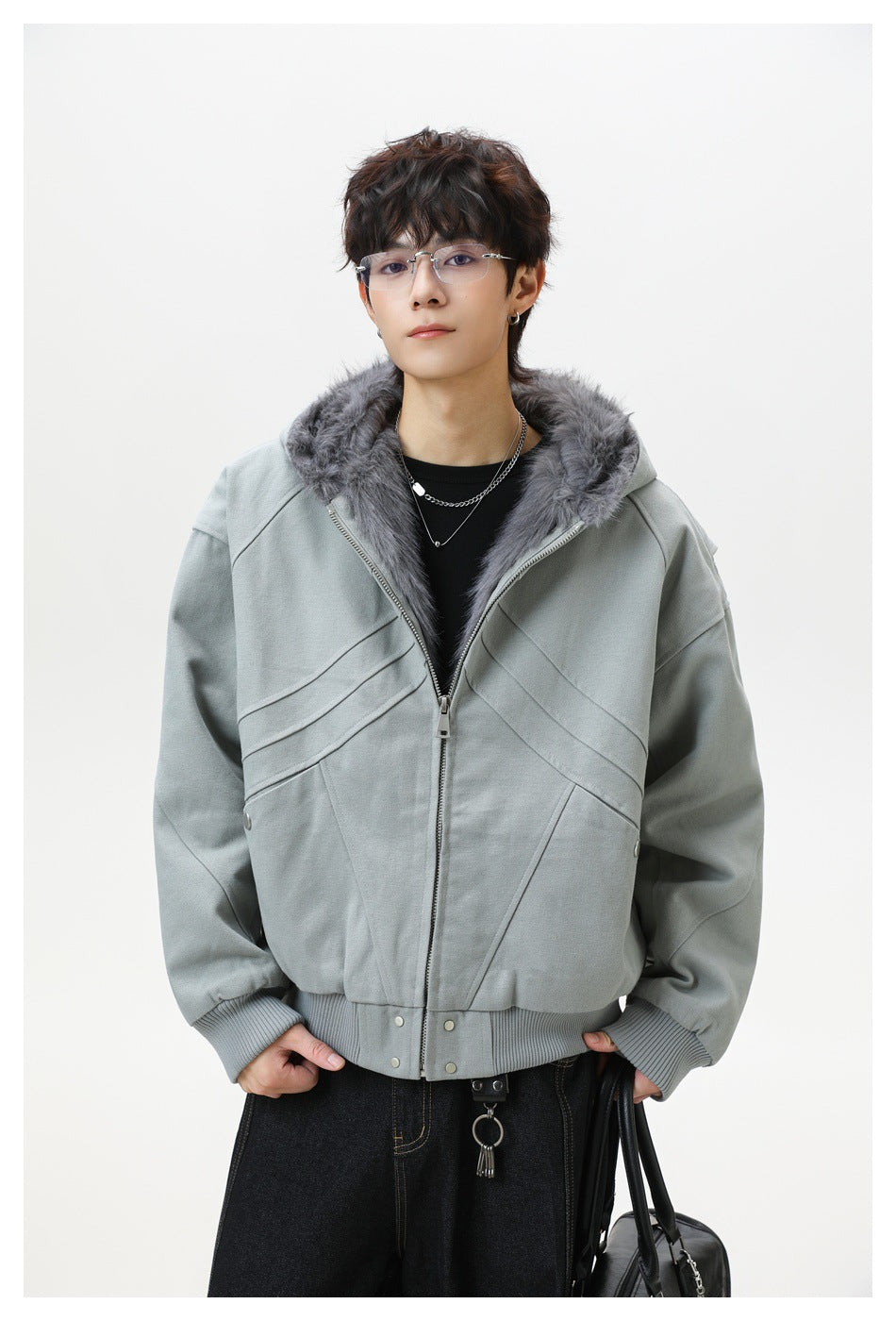 Model wearing a sage-gray zip-front hooded jacket with dark gray faux-fur lining and diagonal seams, shown from the waist up.