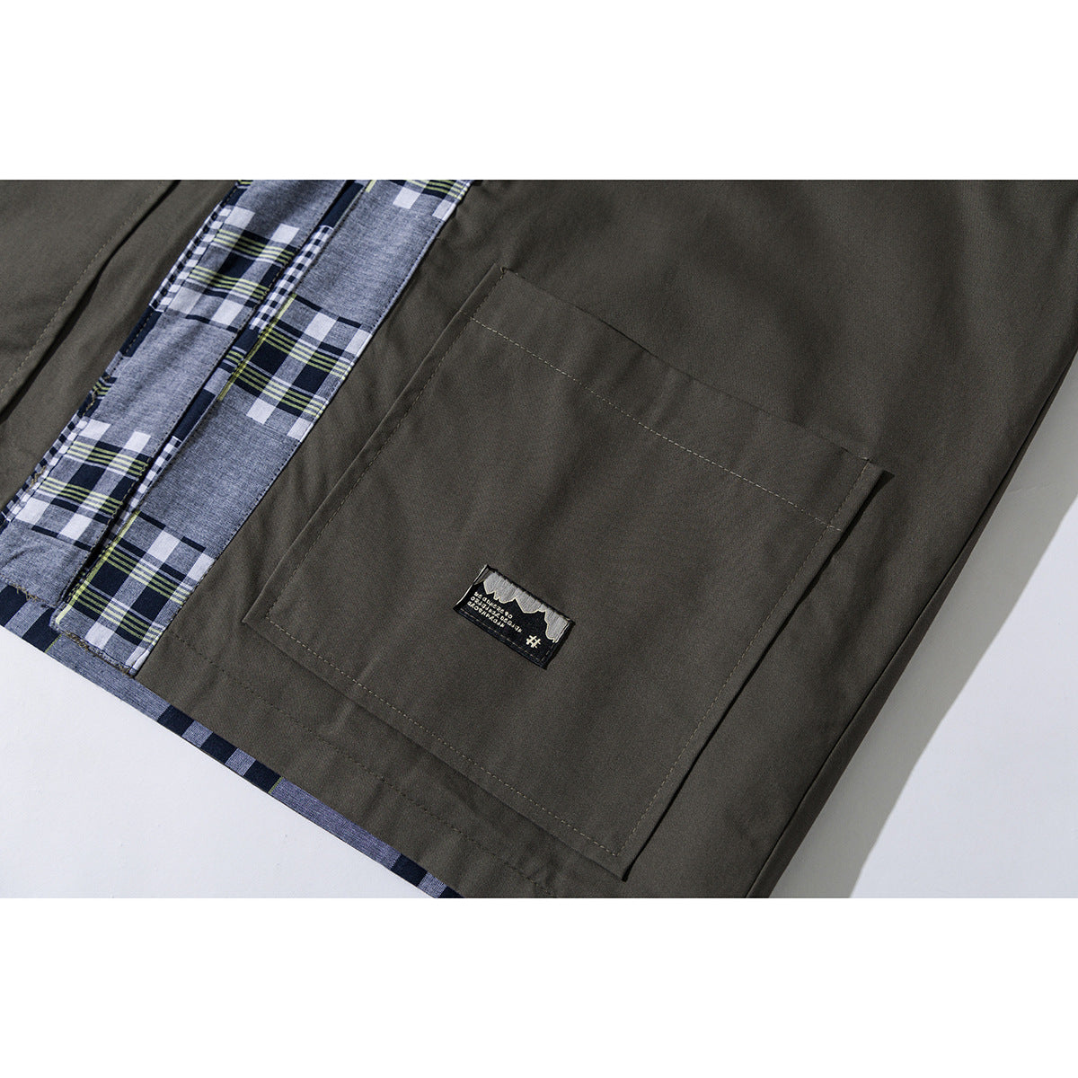 Close-up of an olive jacket's large front pocket beside blue-gray plaid trim and a small sewn label.