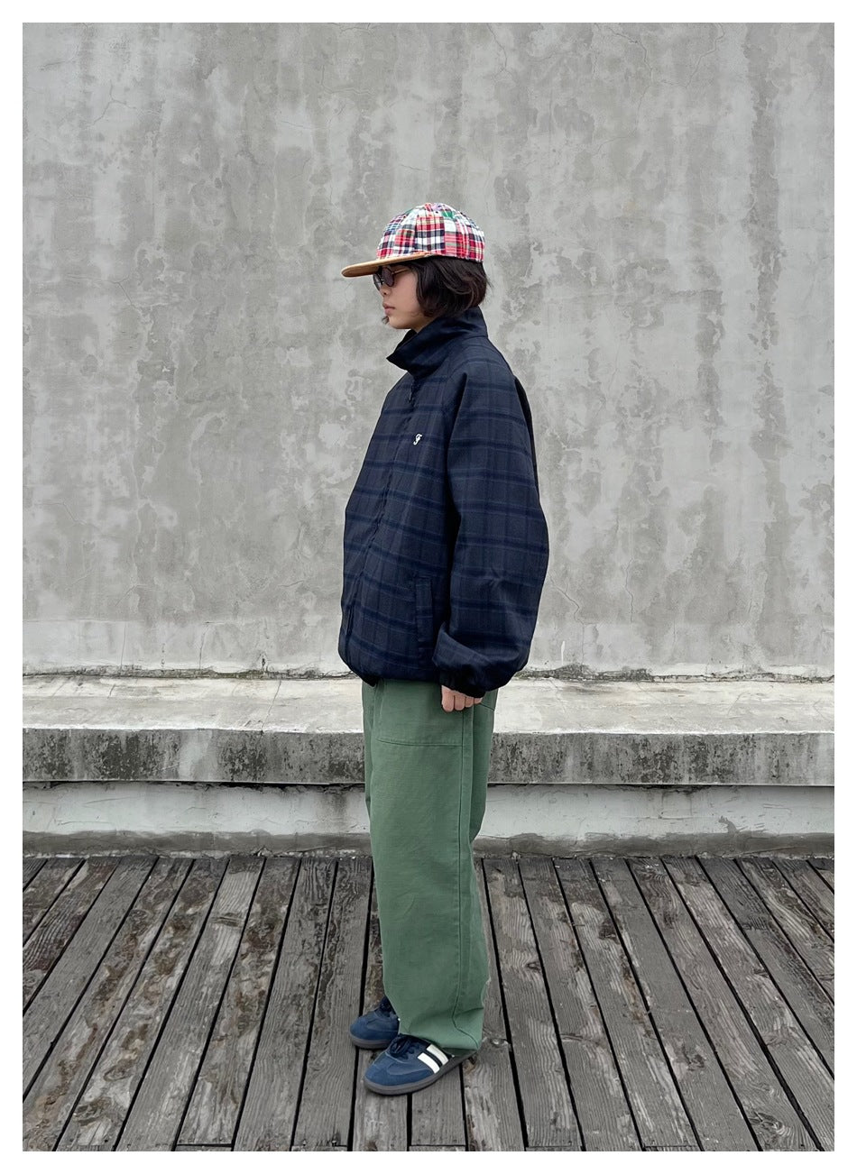 Side view of a person wearing a navy plaid zip jacket with green pants, a patchwork cap and blue sneakers