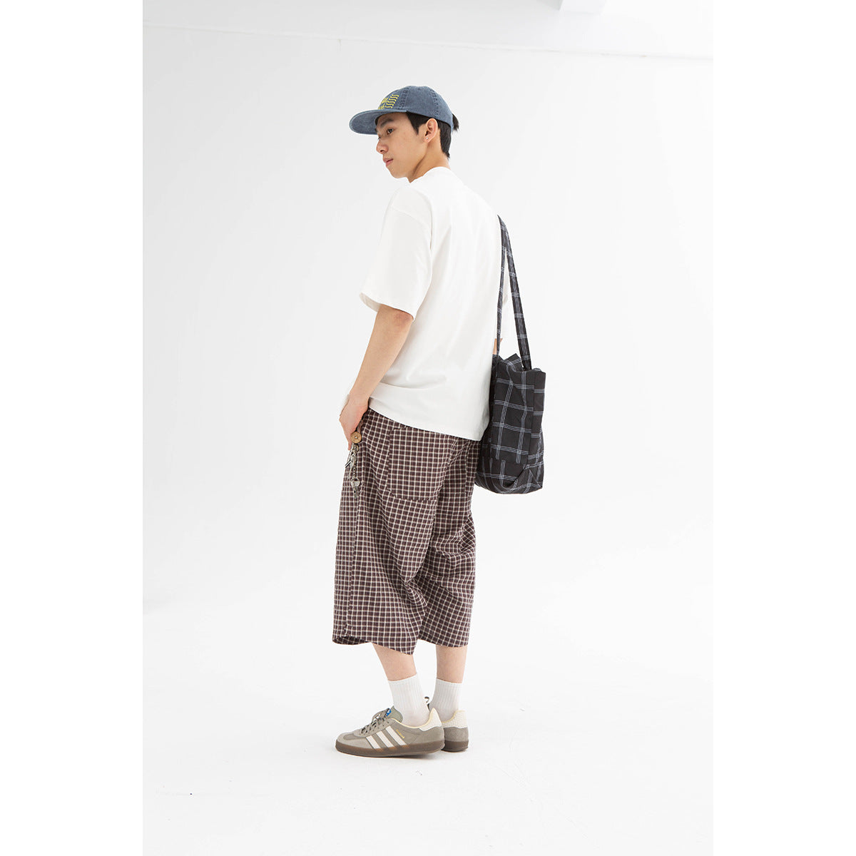 Model wearing a white T-shirt and brown plaid shorts, shown from the back with a shoulder bag.