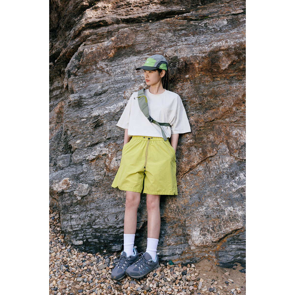 Woman wearing a white pocket T-shirt, lime-green shorts and cap, posing against a rocky backdrop.