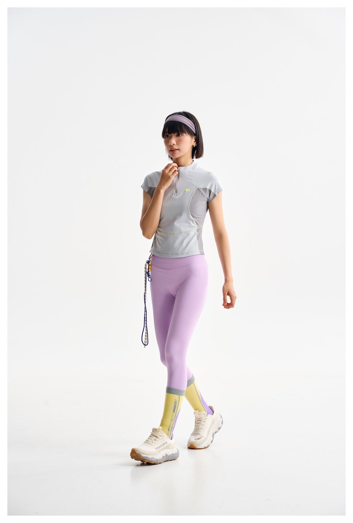 Model wearing a pale gray cap-sleeve quarter-zip top with darker panels and lilac leggings.