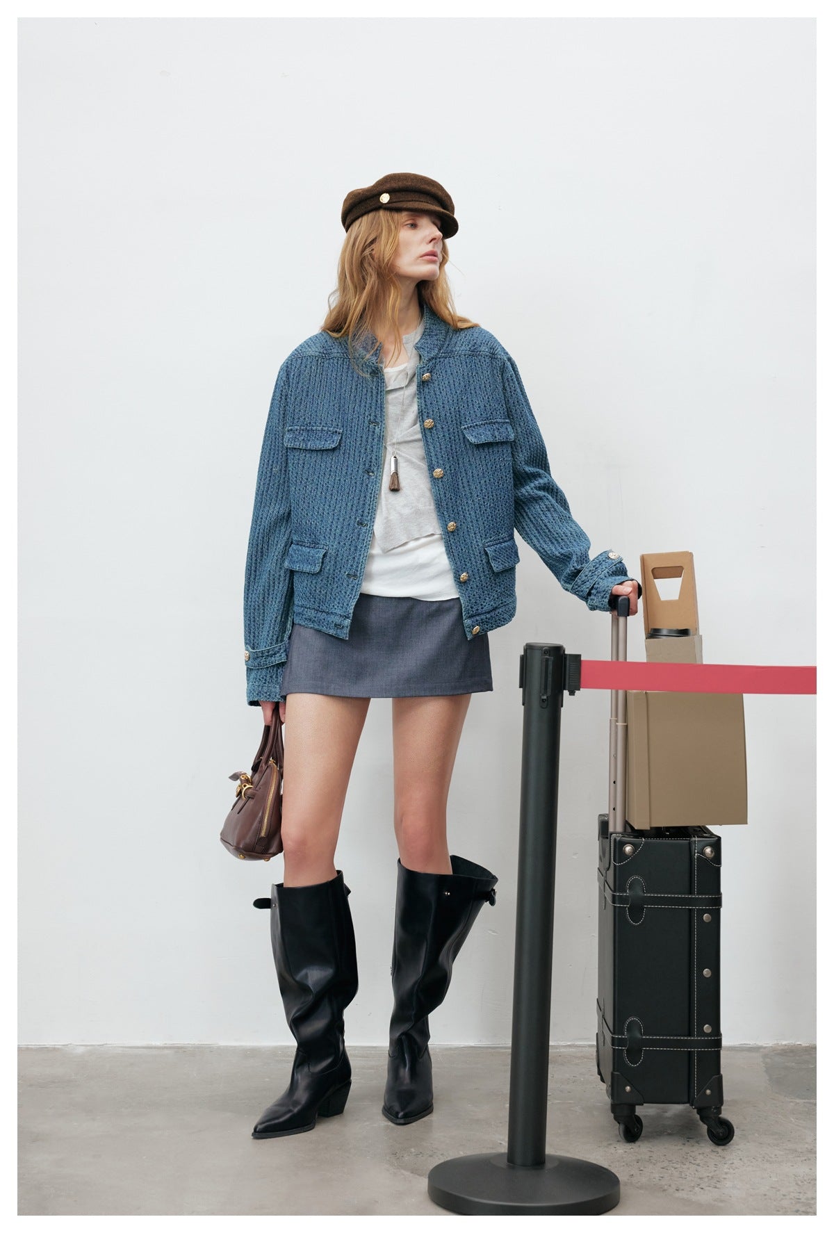 Woman wearing washed blue textured stand-collar jacket with a gray top, mini skirt and tall black boots, beside luggage