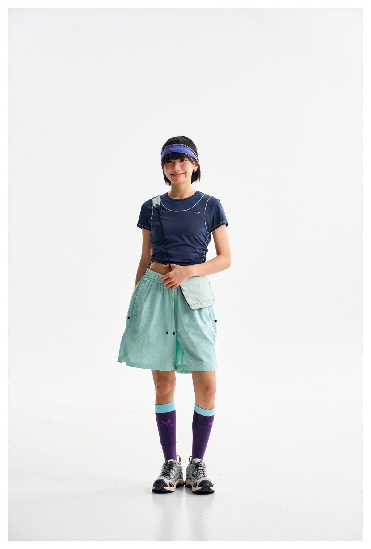 Model wearing a navy fitted tee with pale piping, mint-green shorts, a purple headband, and a small crossbody pouch.