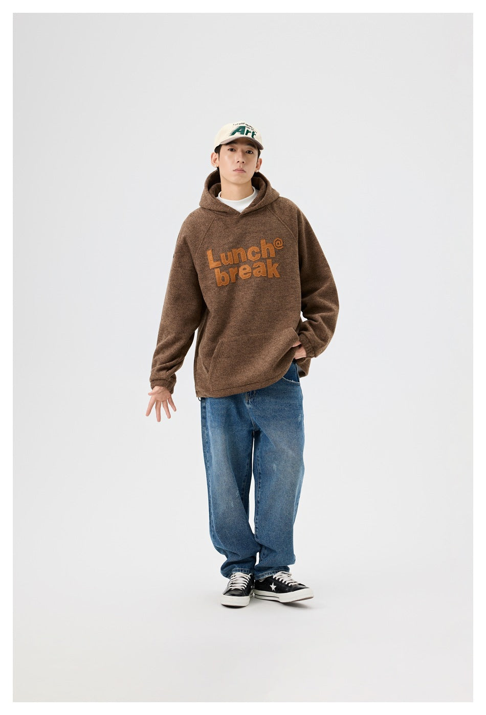 Model wearing a brown hooded pullover with raised orange “Lunch break” lettering and blue jeans.