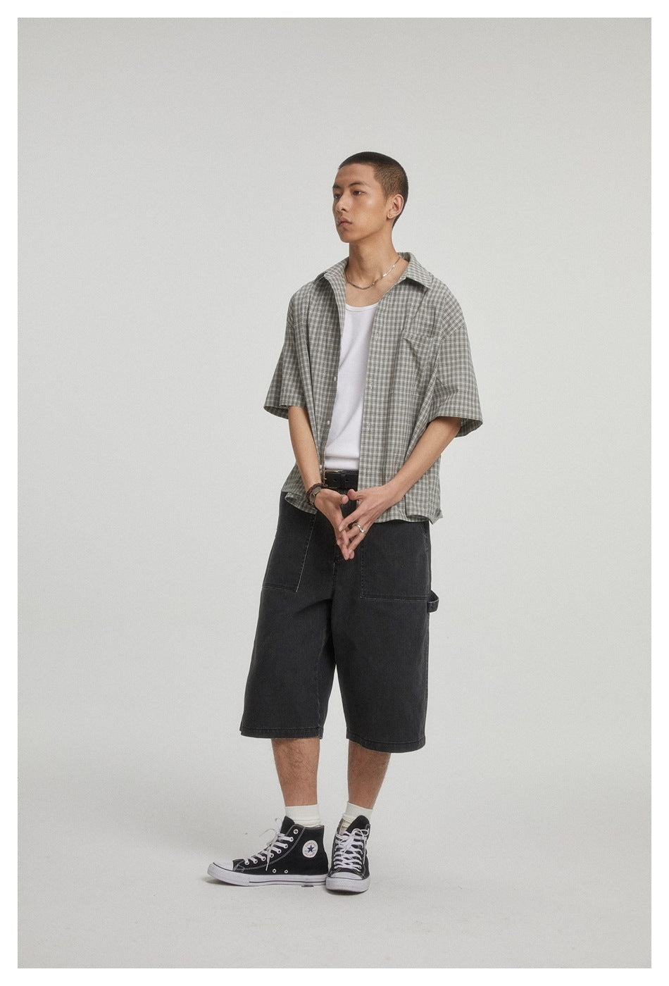 Model wearing an open gray checked short-sleeve shirt over a white tank top with dark shorts.