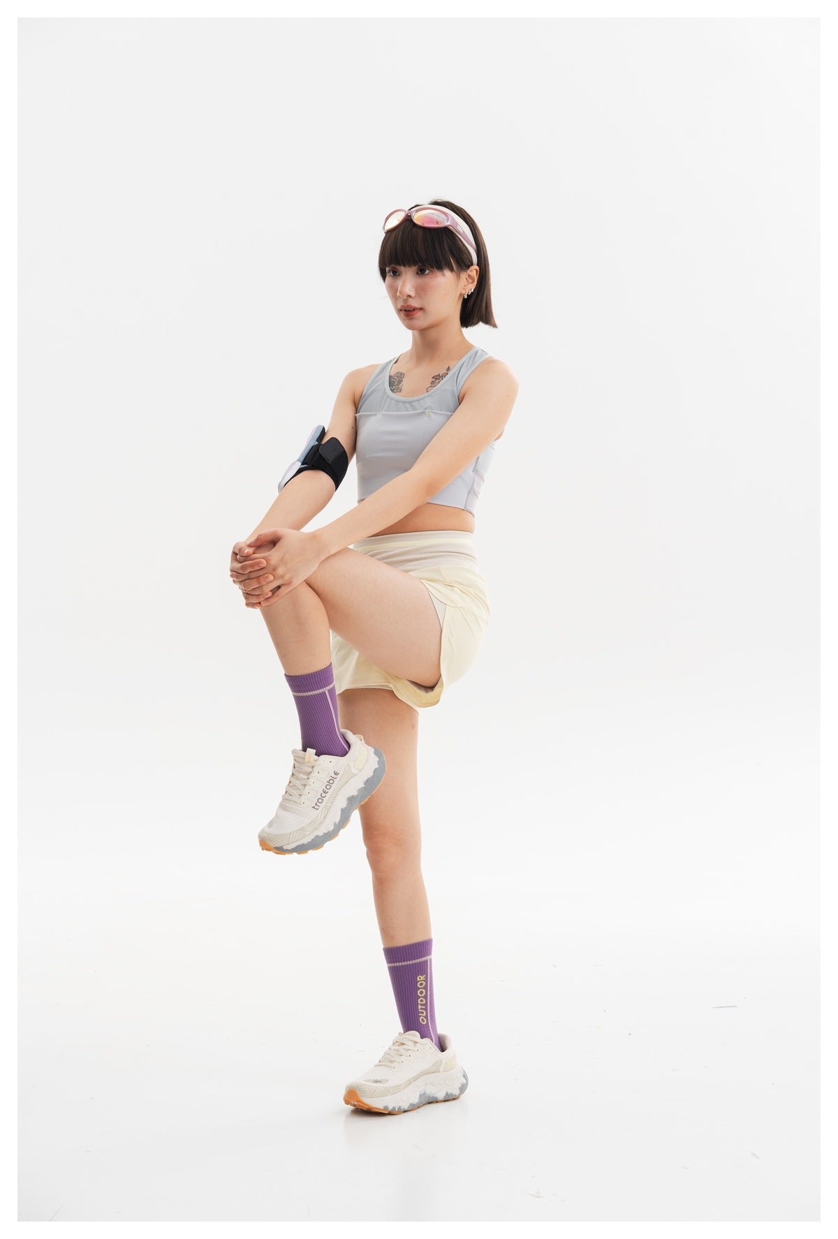 Side-view pose of a model wearing a light gray cropped athletic tank, pale yellow shorts, and a padded arm accessory.