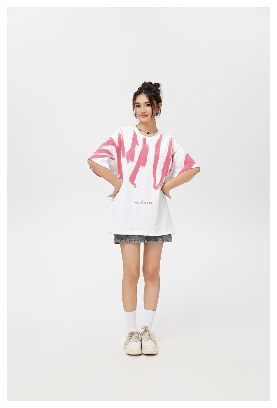 Model facing forward in an oversized white T-shirt with pink brushstroke graphics and gray shorts.