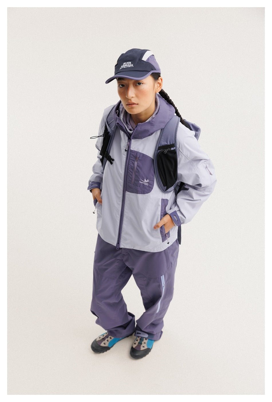 Model wears a light lavender and purple hooded jacket with matching pants, a cap, and a black backpack.