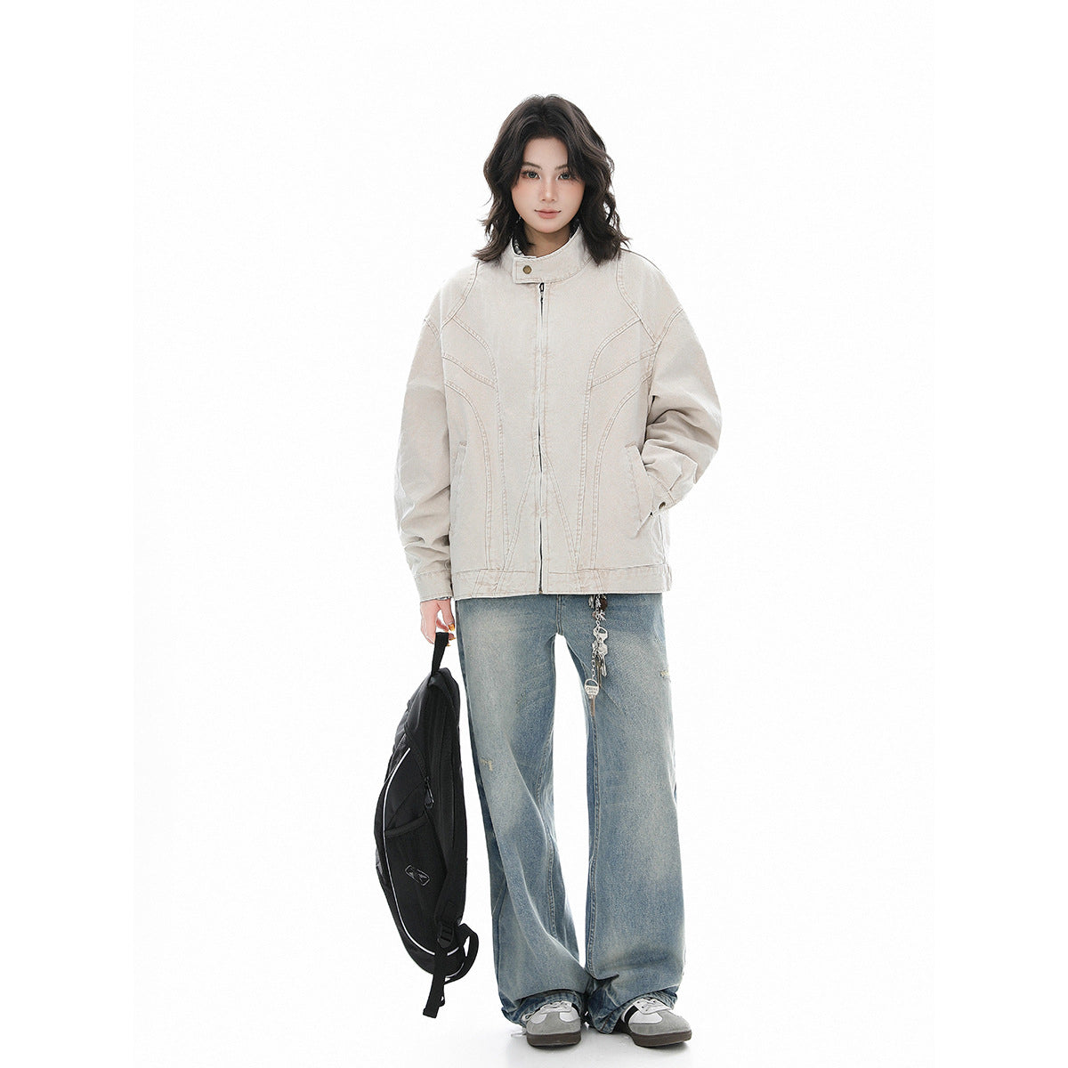 Model wearing a light beige stand-collar jacket with curved seams and blue wide-leg jeans, carrying a black backpack.