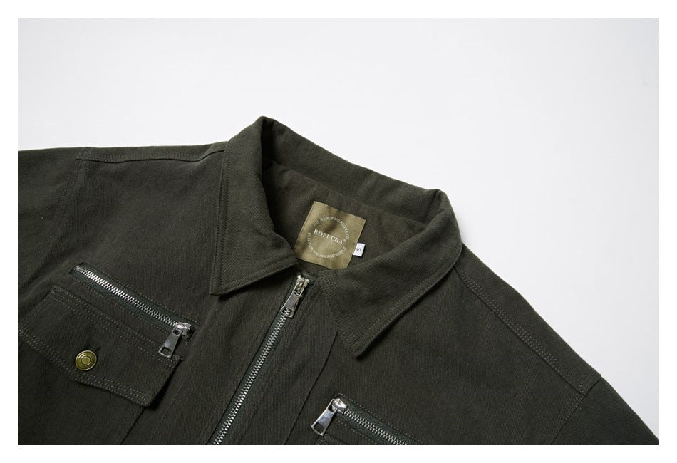 Close-up of a dark olive workwear jacket with a pointed collar, central zipper, and zippered flap pockets.
