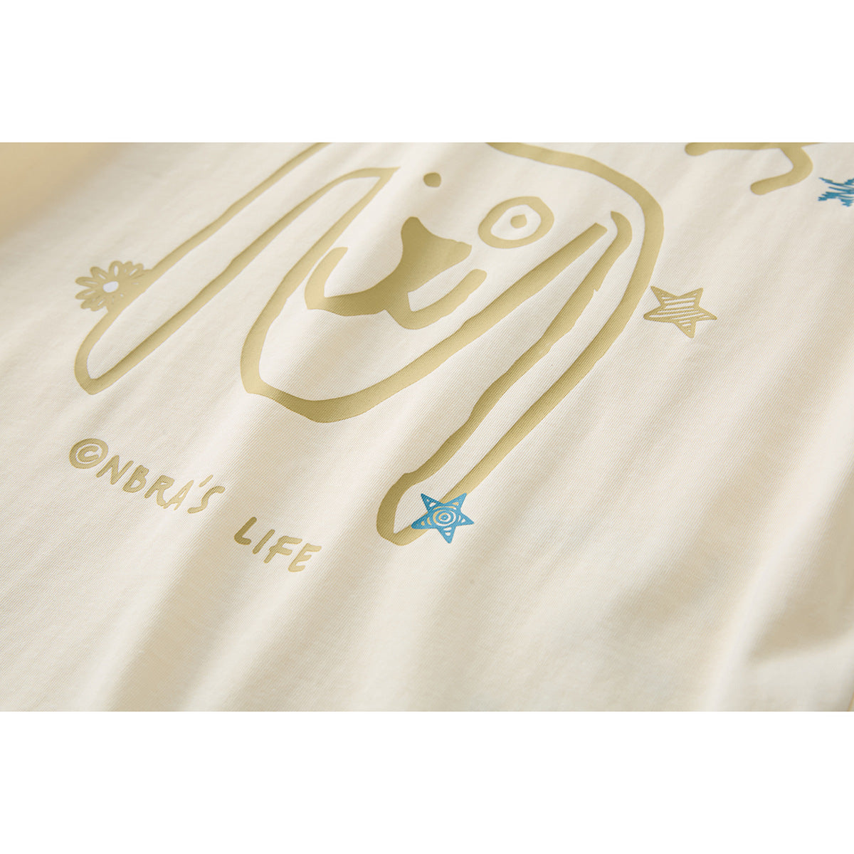 Close-up of a cream T-shirt with a playful face graphic, stars, and English lettering in gold and blue.