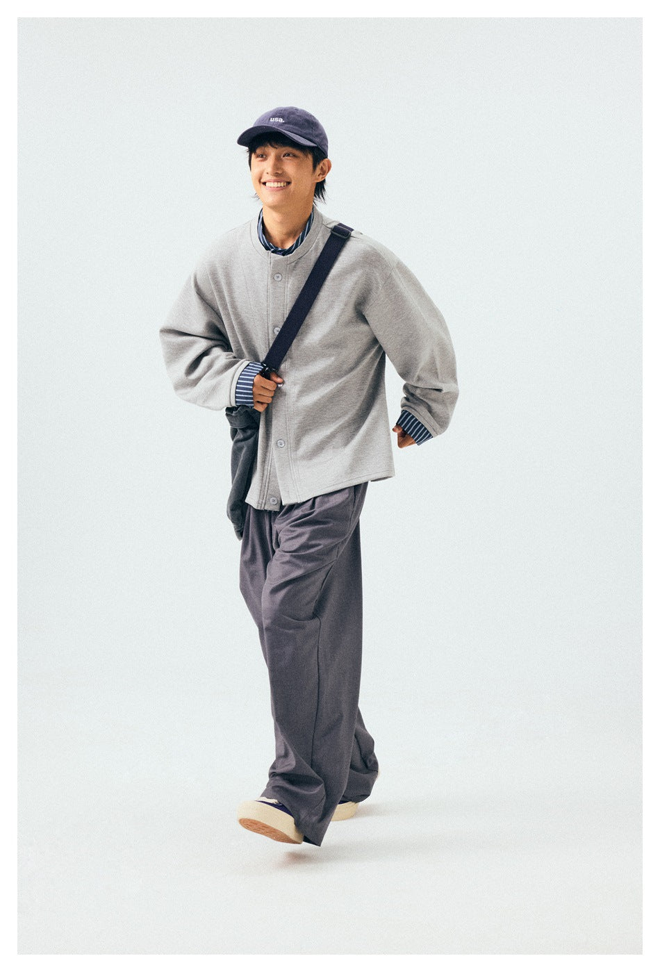 Full-length model wearing loose gray trousers, a matching button-front jacket, and a navy cap.