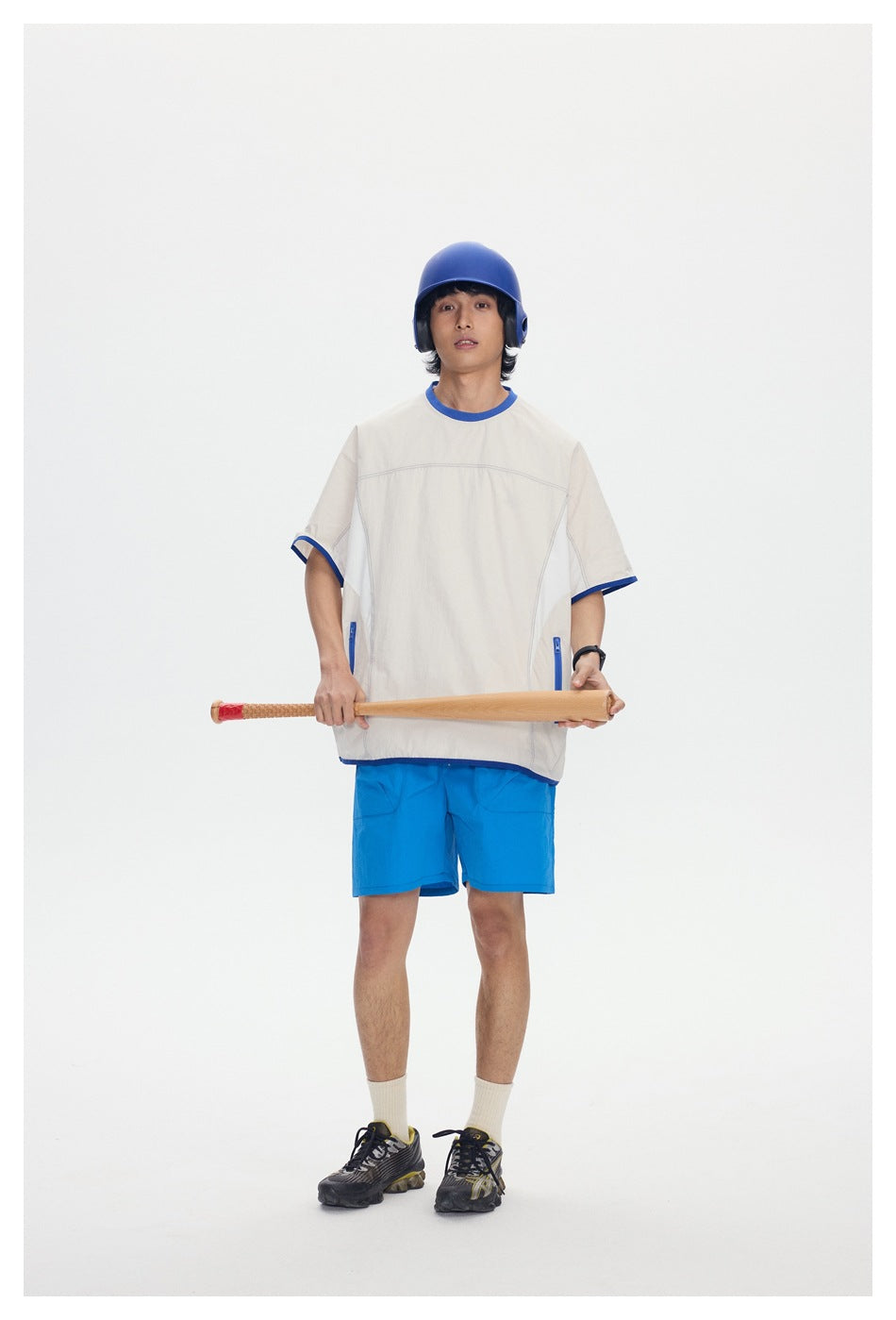 Full-length model wearing an off-white short-sleeve top with blue trim and bright blue shorts, holding a bat.
