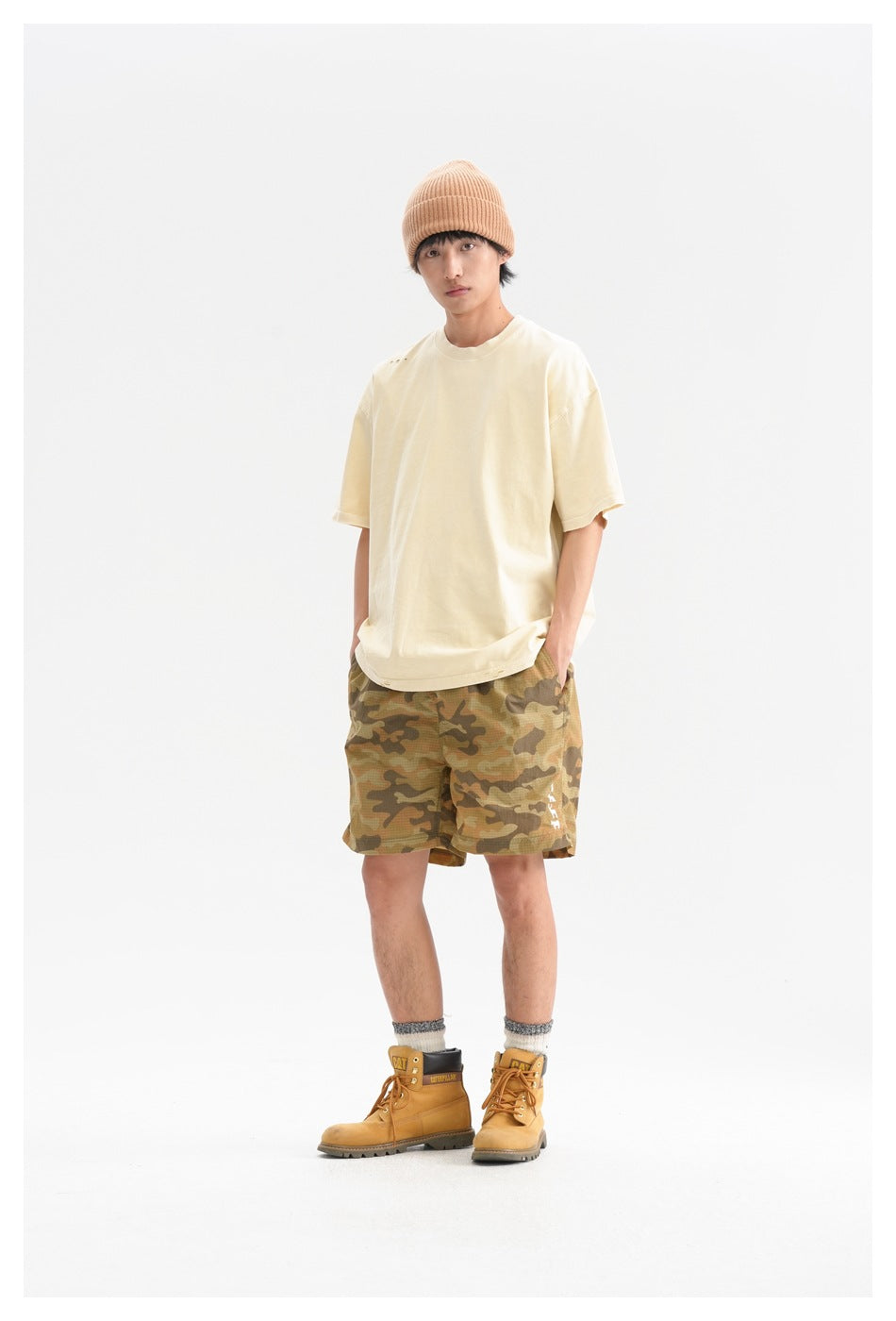 Model wearing a light yellow oversized T-shirt with camo shorts, beanie and tan boots, standing front view