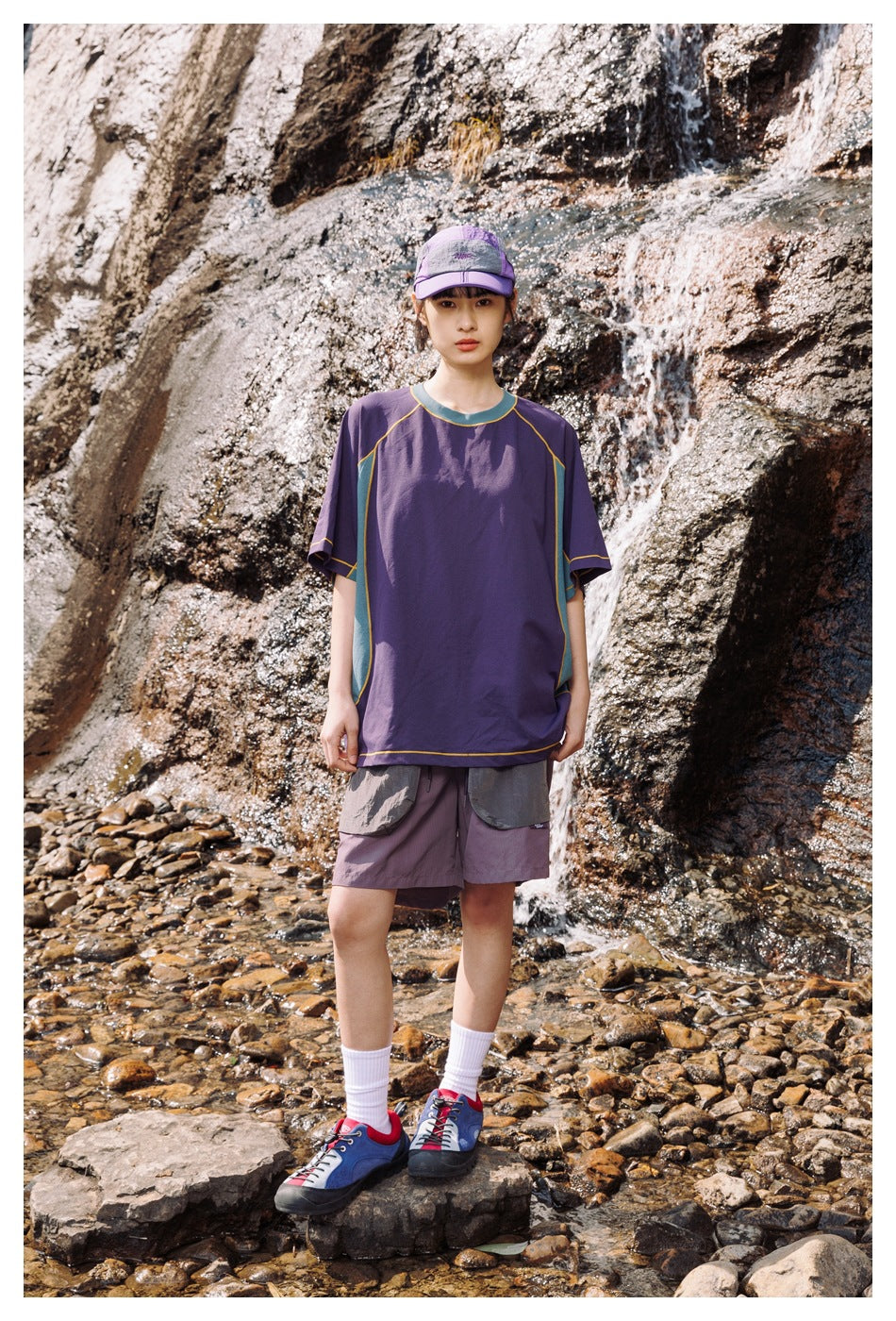 Model standing by a waterfall wearing the purple T-shirt with teal side panels, gray shorts, a cap and sneakers