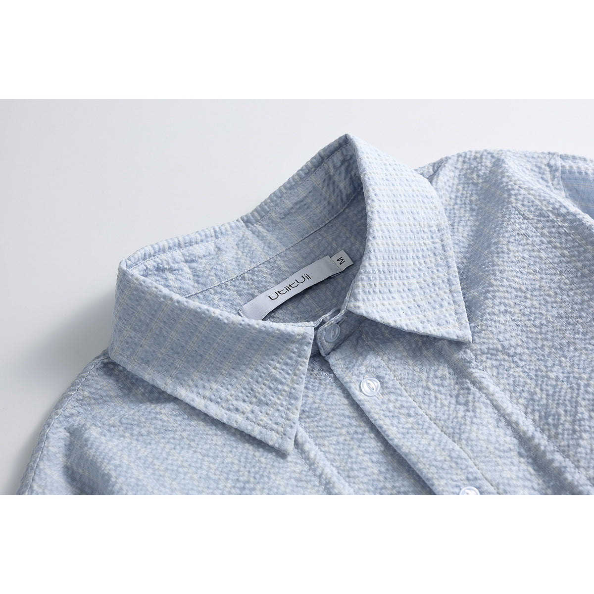 Close-up of a pale blue textured shirt collar, white buttons, and sewn-in neck label.