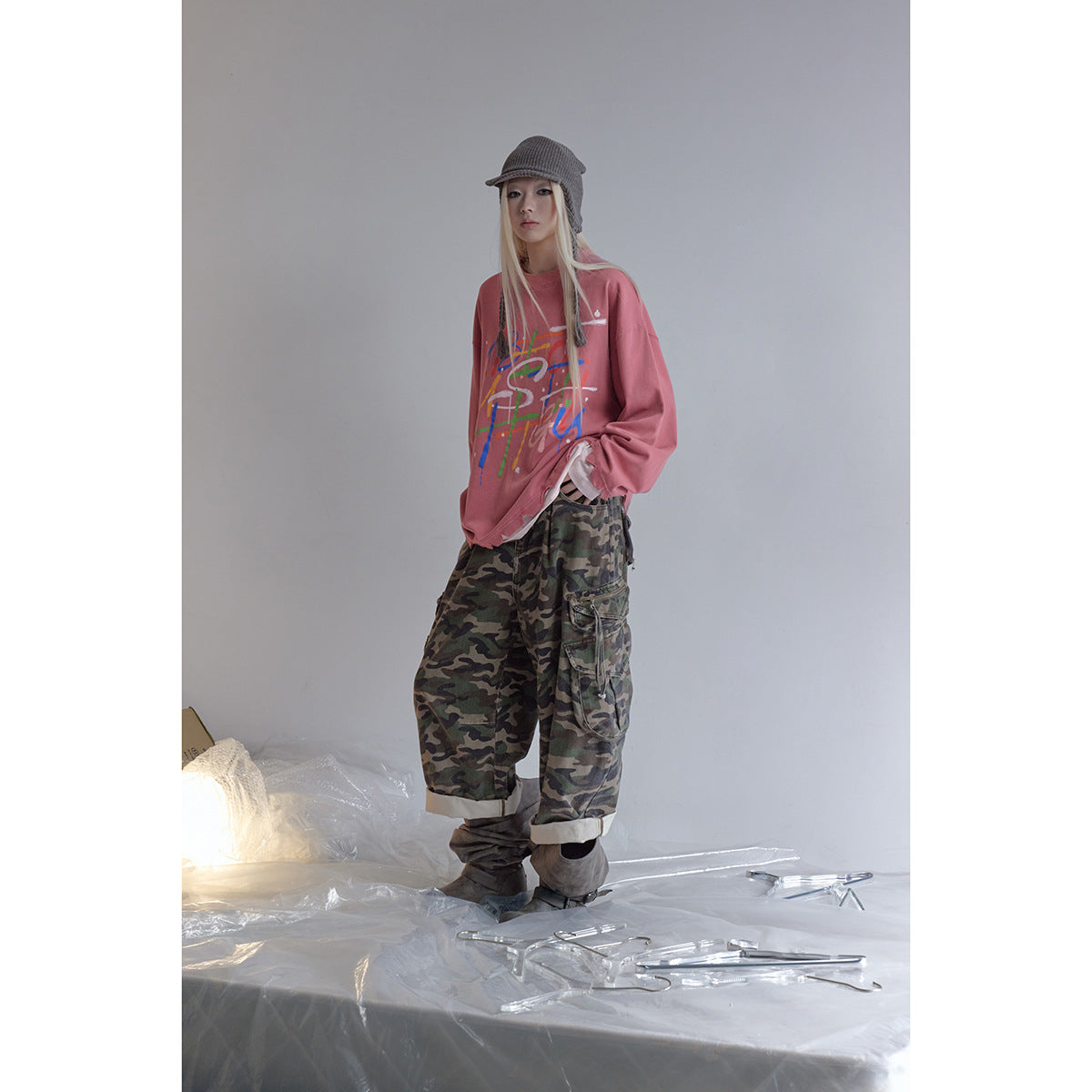 Model wearing a dusty rose oversized graphic sweatshirt with camouflage cargo pants and a gray knit cap.