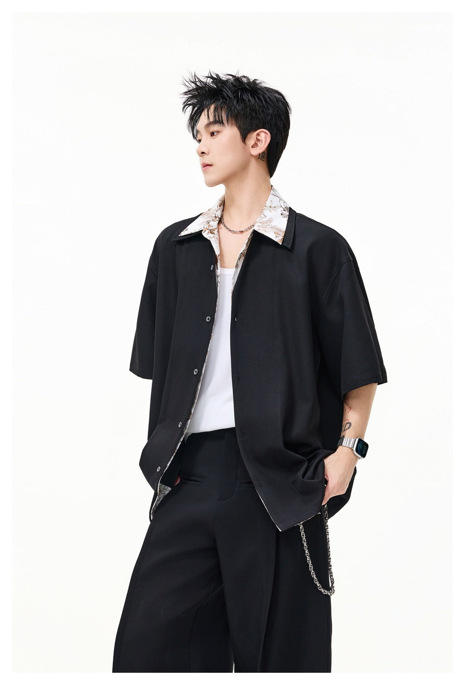 Model wearing a loose black short-sleeve button-up with a light floral collar, white tank, and black trousers.