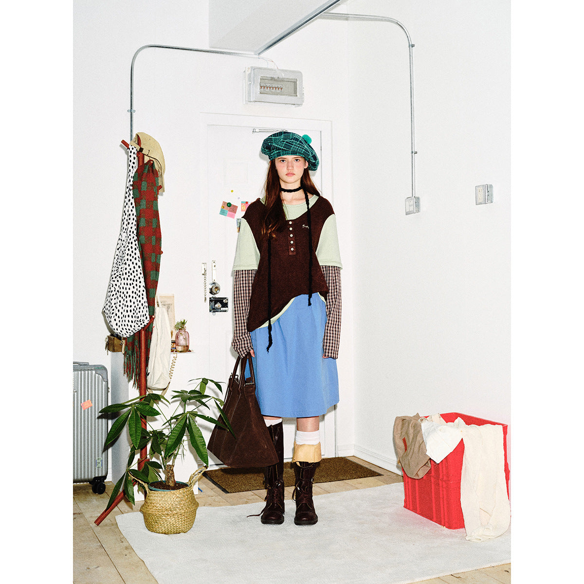 Front view of a model wearing a green plaid beret, layered sage and burgundy top, blue skirt, and dark boots.