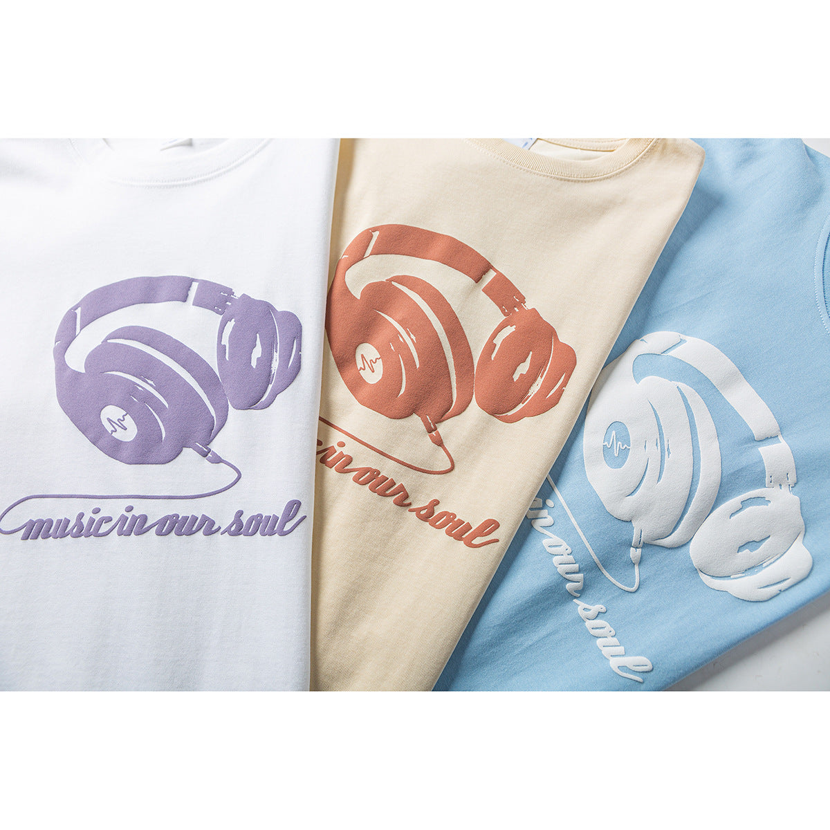 Three folded T-shirts in white, cream, and light blue, each printed with headphones and “Music in our soul.”