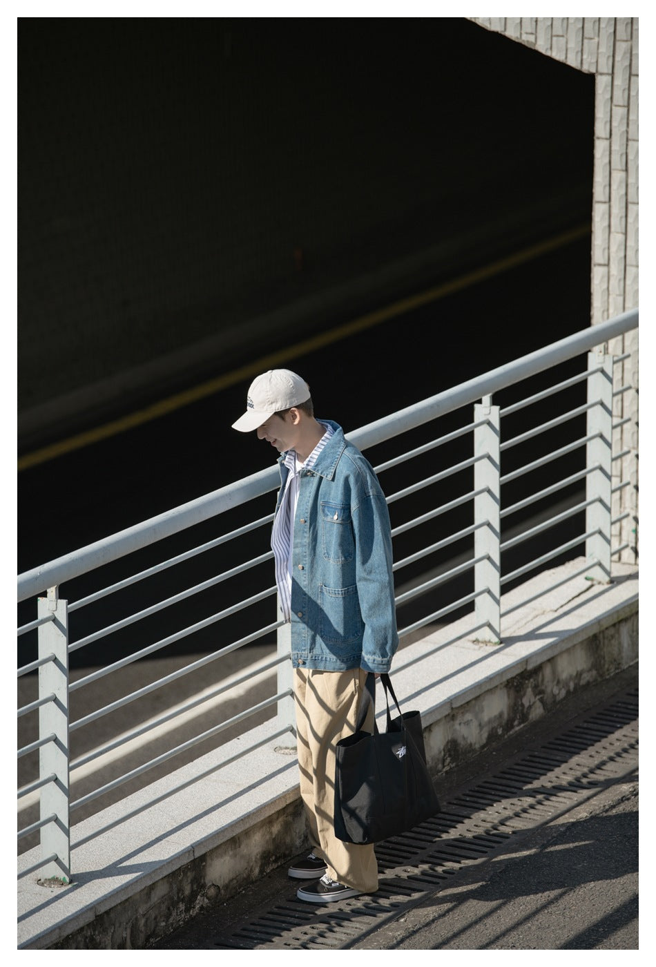 Side view of a model wearing a blue denim jacket and tan trousers, carrying a black tote outdoors.