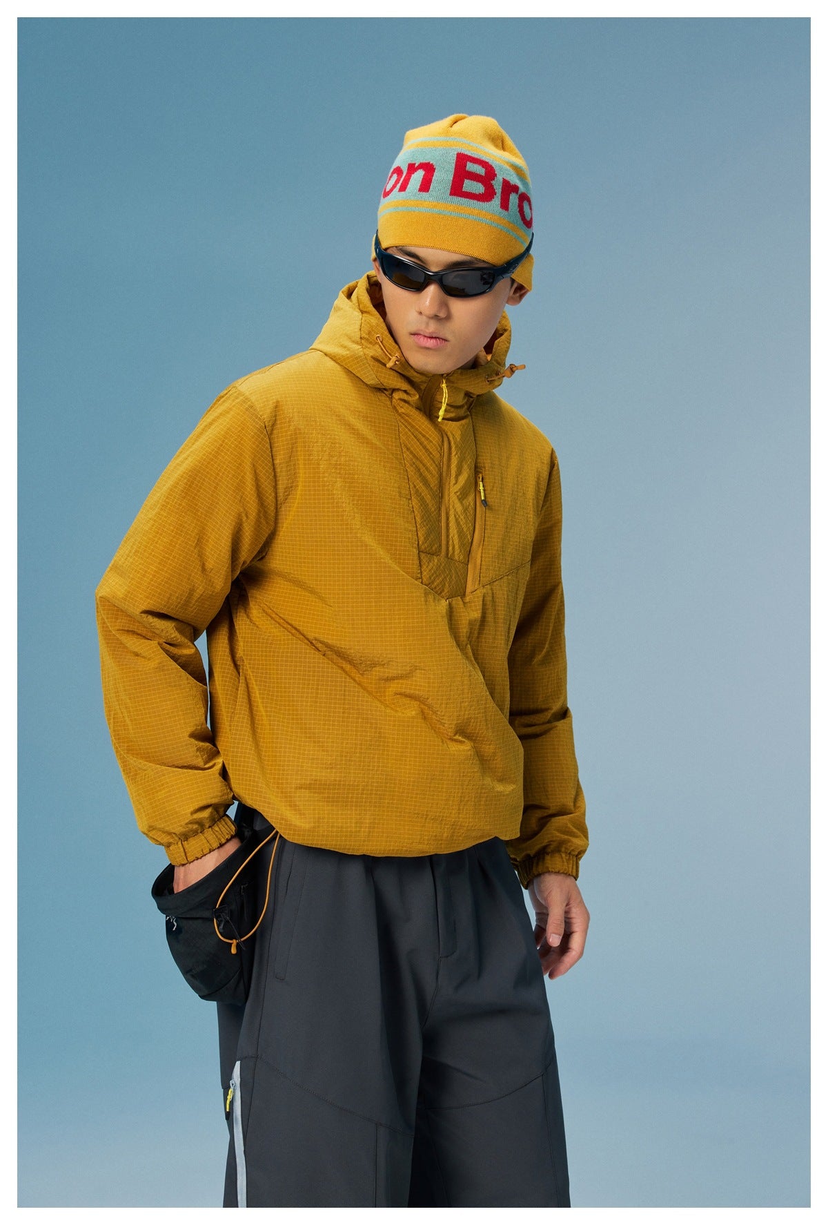Model wearing a mustard grid-pattern hooded pullover with dark pants, shown from the front.