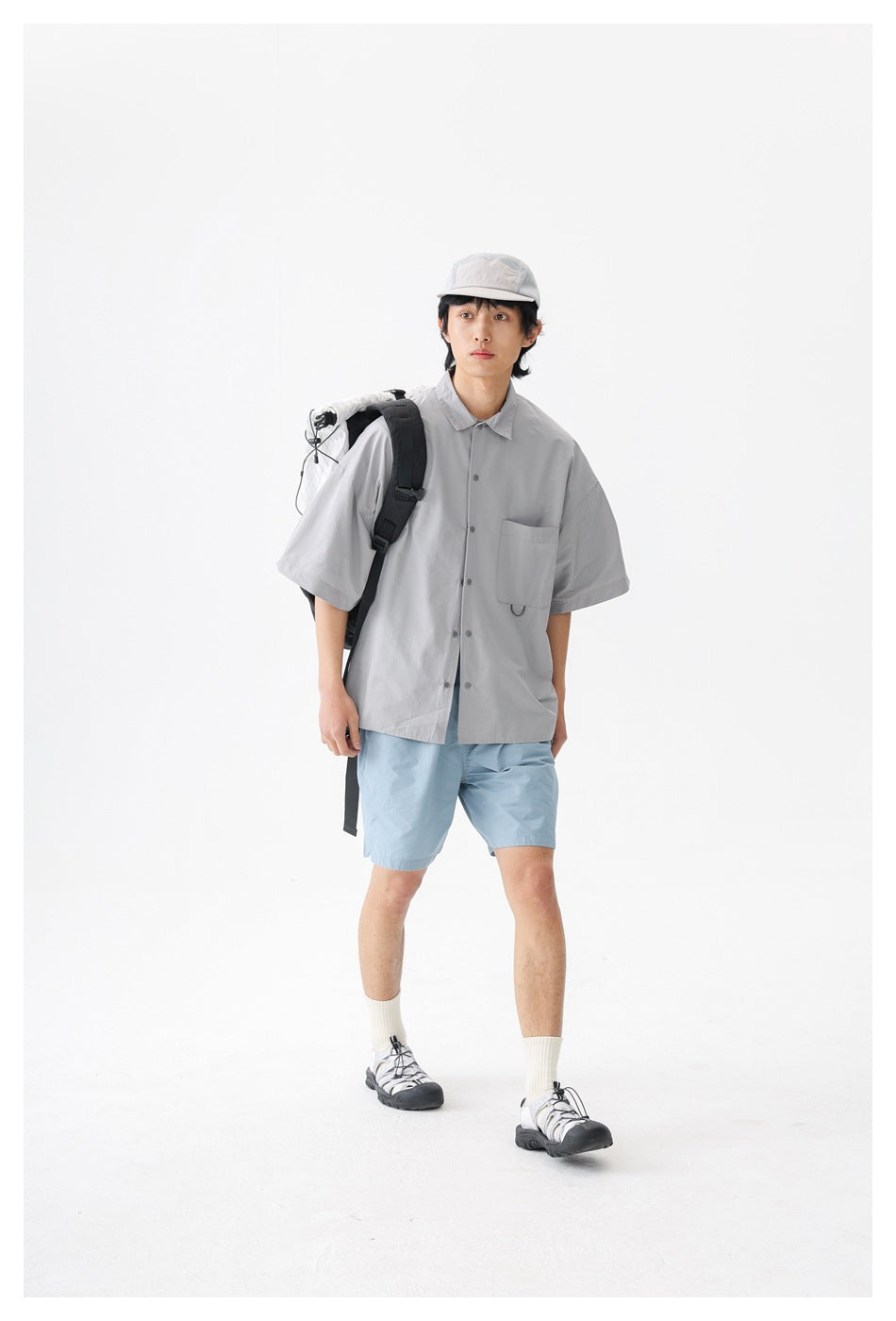 Model wearing a light gray short-sleeve button-up shirt and pale blue shorts with a backpack over one shoulder.