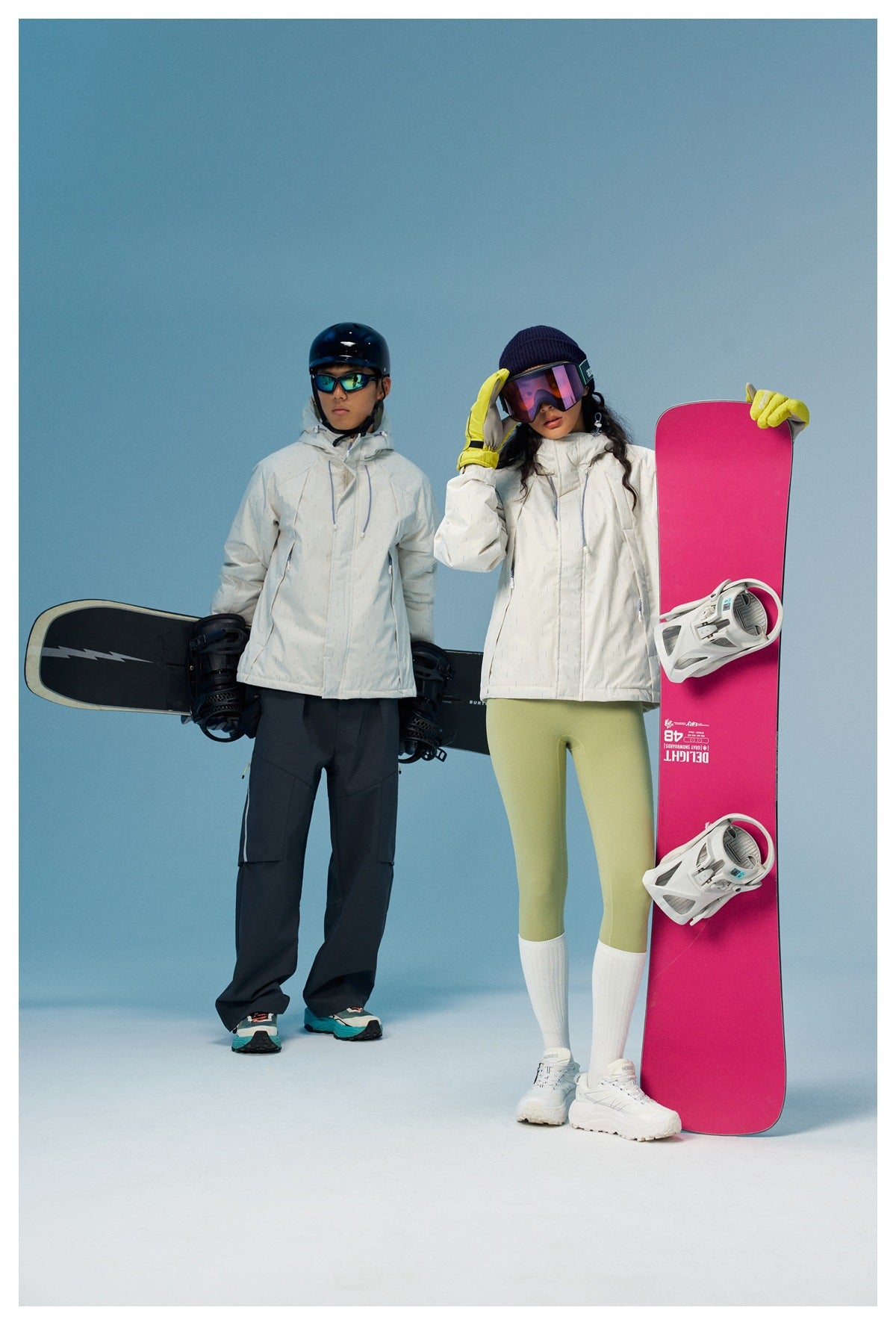 Two models in off-white hooded jackets holding snowboards, woman adjusting her goggles, blue studio background