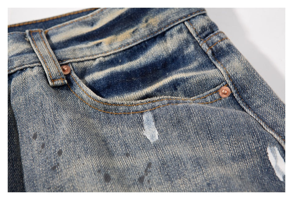 Close-up of a front pocket on faded blue jeans, with copper rivets and distressed paint-like marks.