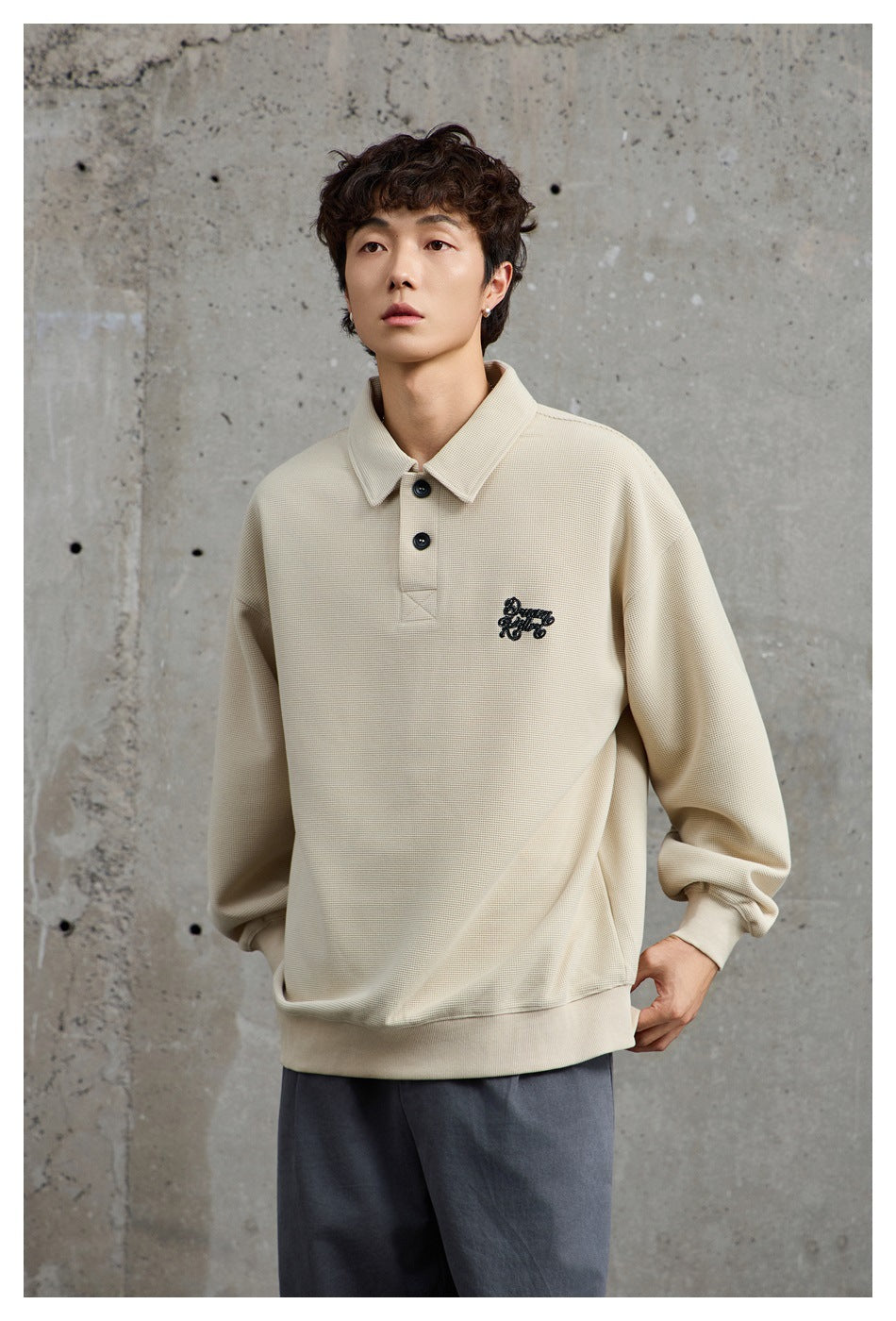 Model wearing a light beige collared pullover with a two-button placket, dark chest embroidery, and ribbed cuffs and hem.