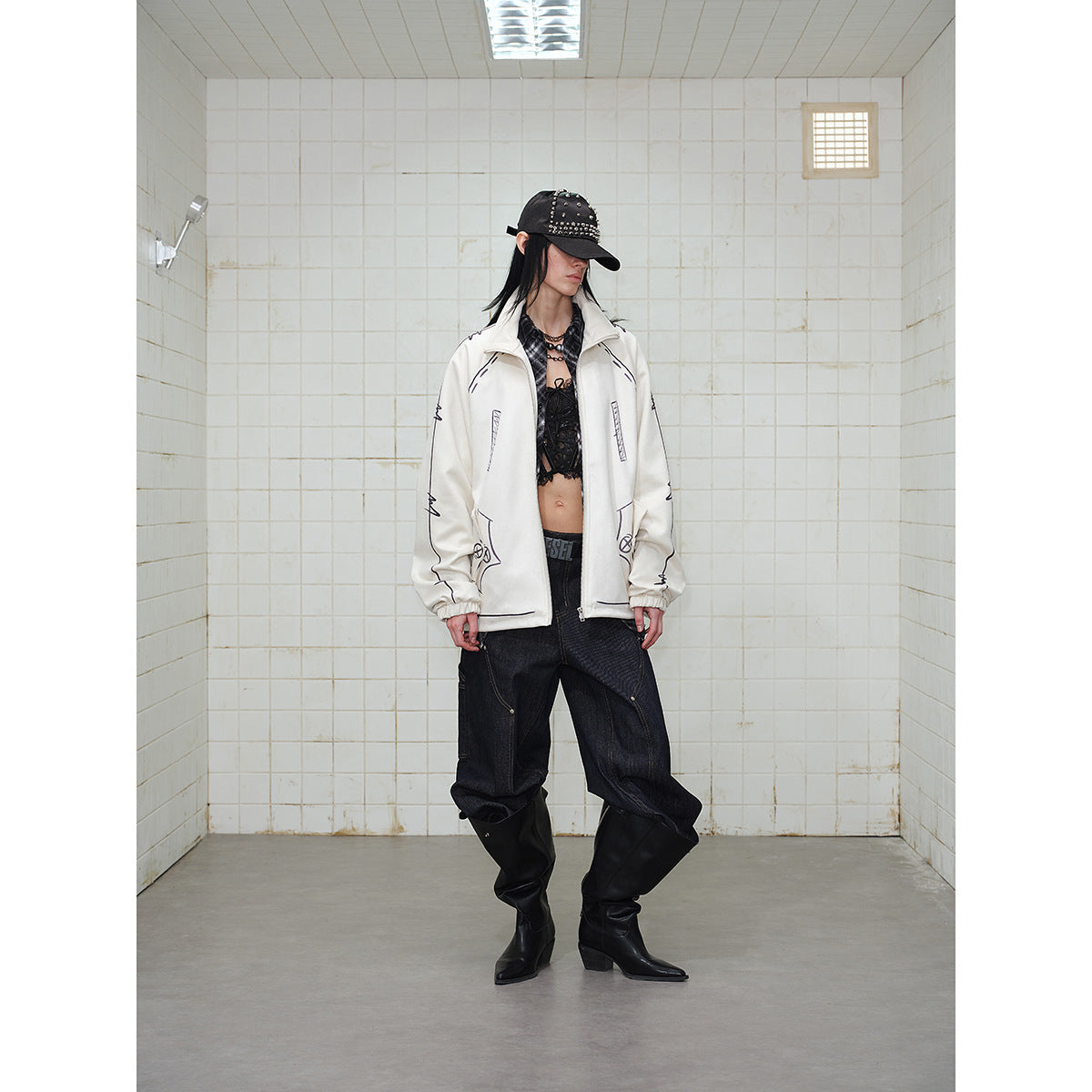 Front view of a model wearing a cream graphic zip jacket with dark baggy jeans and a studded black cap.