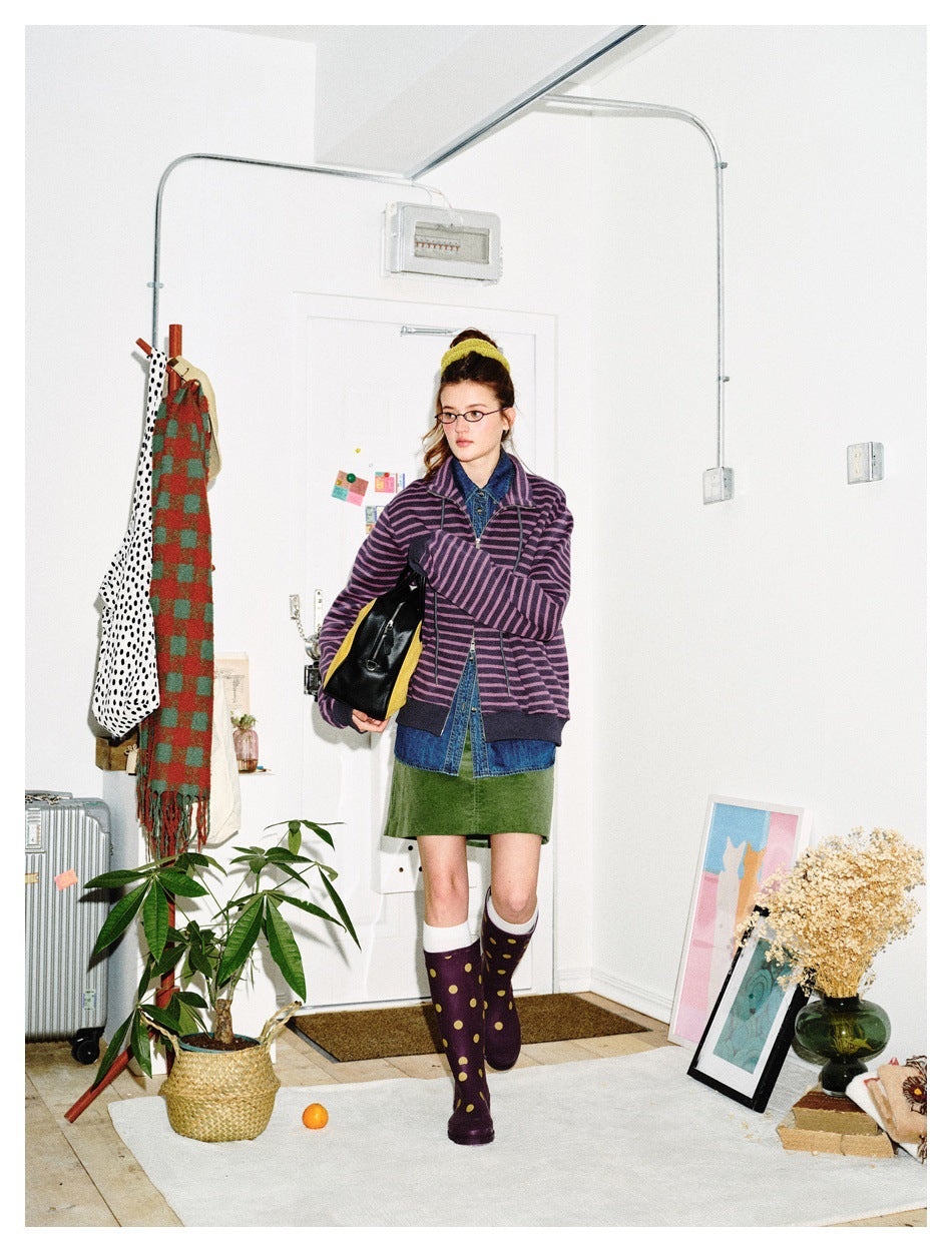 Front view of a model in a purple-and-black striped zip jacket, denim shirt, green skirt, and polka-dot boots.