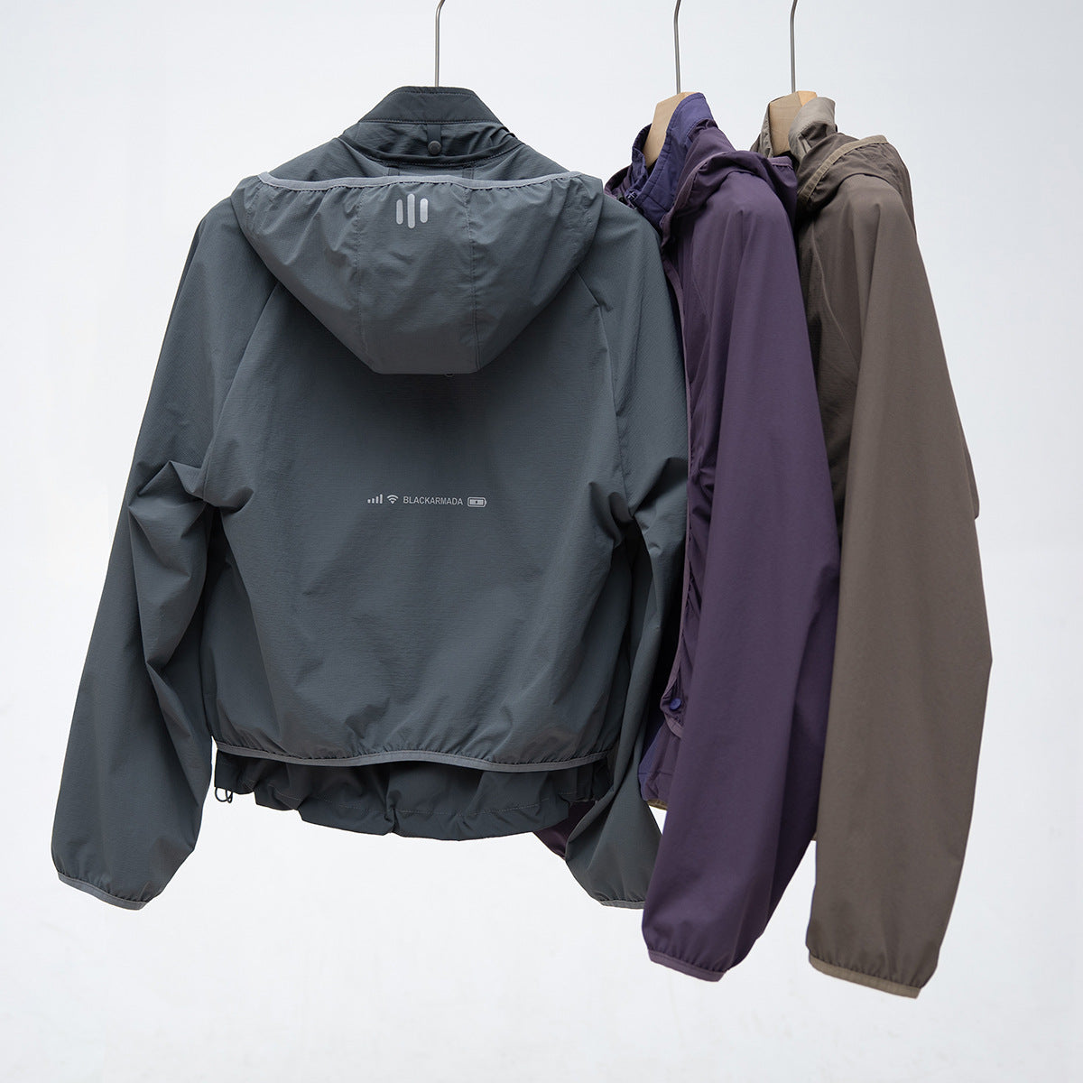 Three hooded jackets hanging back-facing in charcoal, purple, and taupe; the charcoal jacket has a reflective back detail.