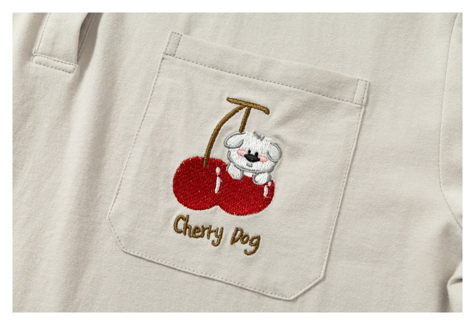 Close-up of a cream shirt pocket embroidered with a white dog, red cherries, and the words “Cherry Dog.”