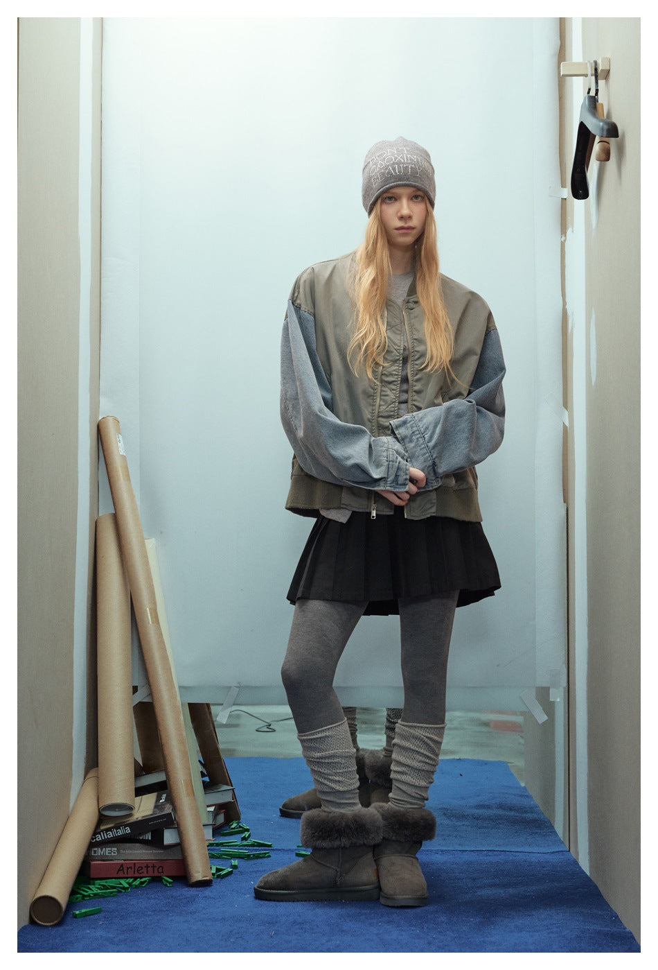 Model wearing an oversized olive-gray bomber jacket with denim sleeves, styled with a beanie, black pleated skirt and boots