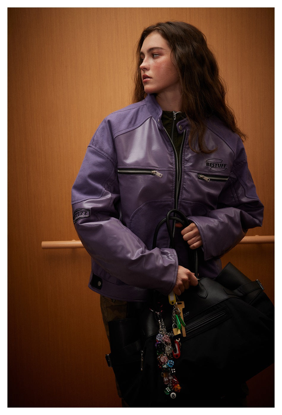 Model wearing a purple paneled zip jacket with chest zip pockets, holding a black bag.