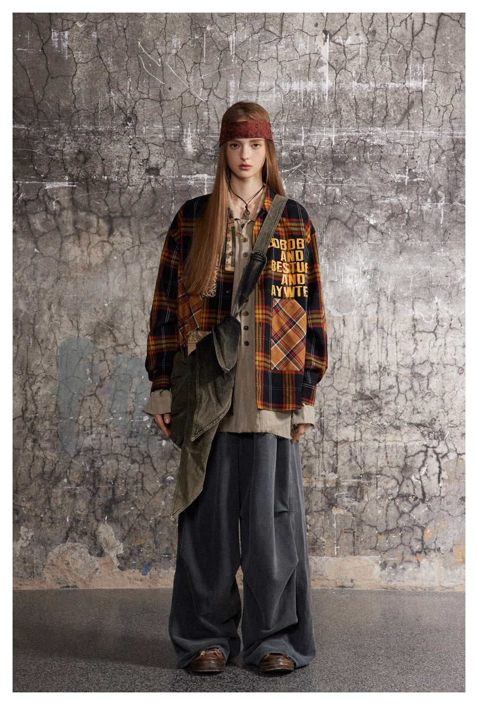 Model wearing the plaid patchwork shirt open over a beige shirt with wide gray pants against a cracked wall