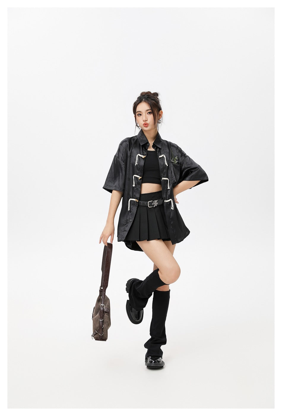 Model in the black rope-closure shirt styled open with a black crop top, pleated mini skirt and a brown shoulder bag