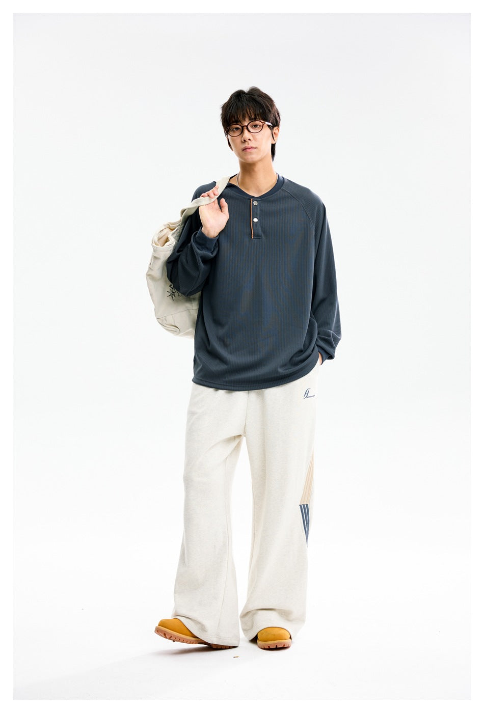 Model wearing a slate-blue long-sleeve Henley shirt and loose cream pants, carrying a light-colored shoulder bag.