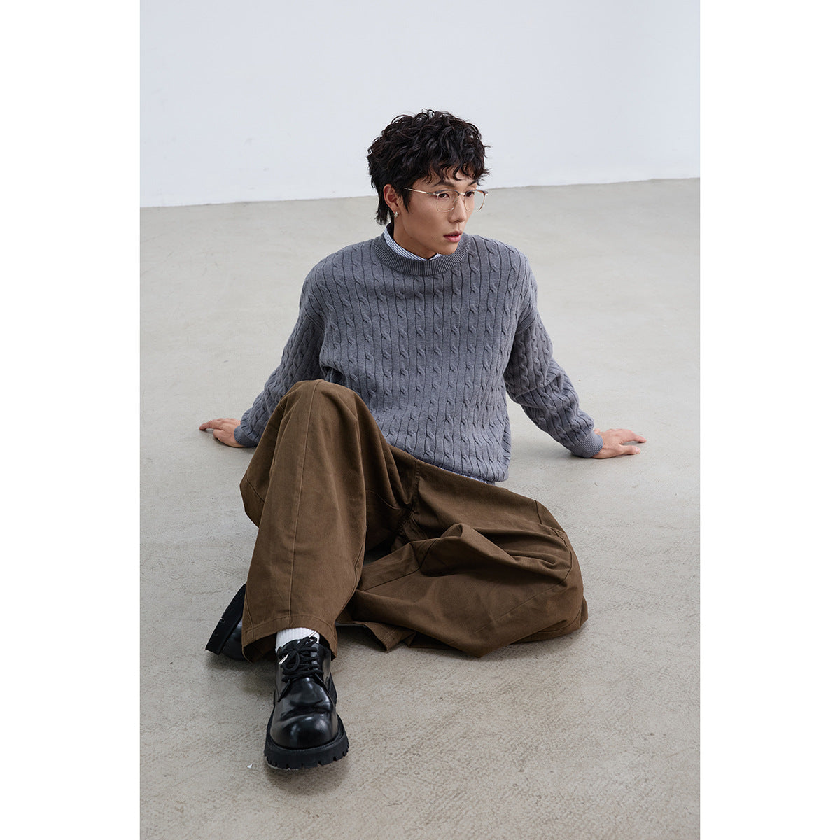 Seated model wearing a gray cable-knit crewneck sweater over a striped shirt with brown trousers.