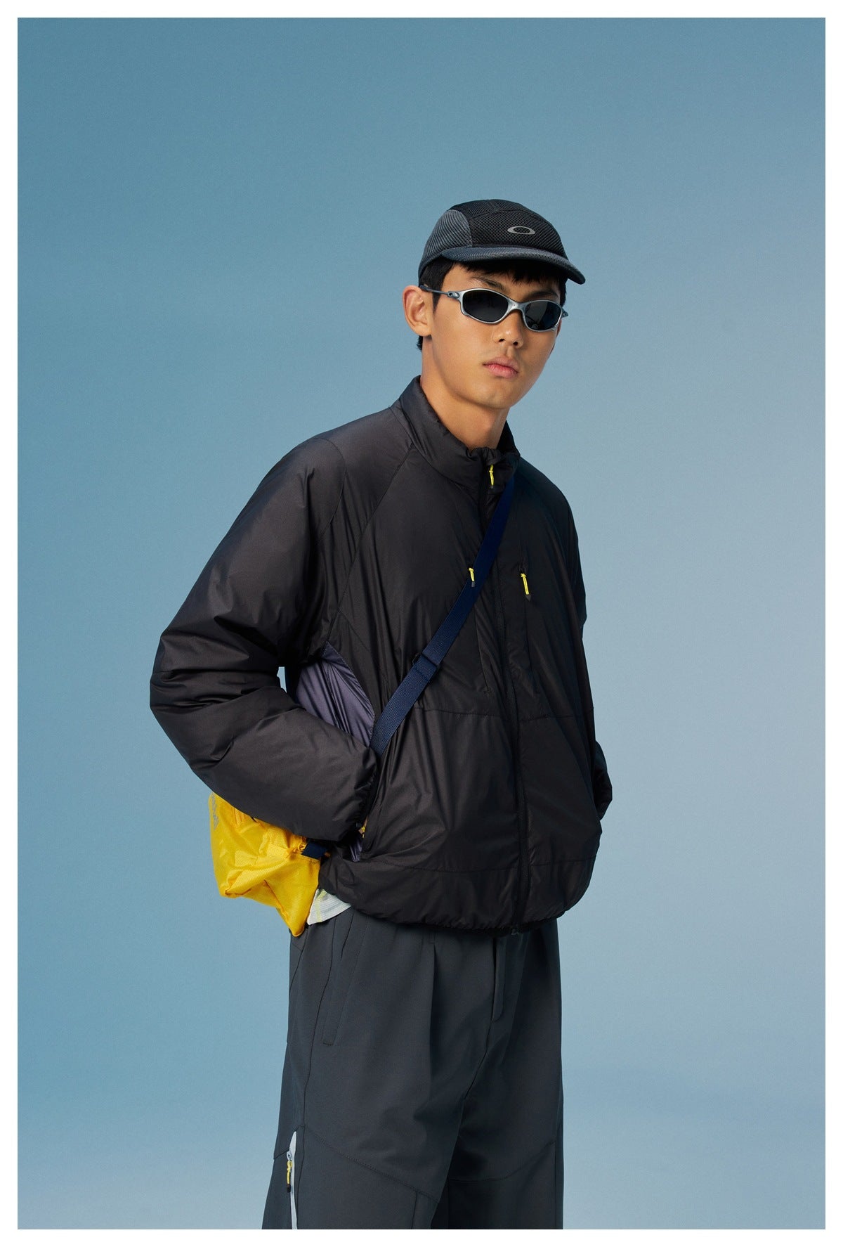 Man wearing a black high-collar zip jacket with yellow zipper pulls and a yellow crossbody bag.